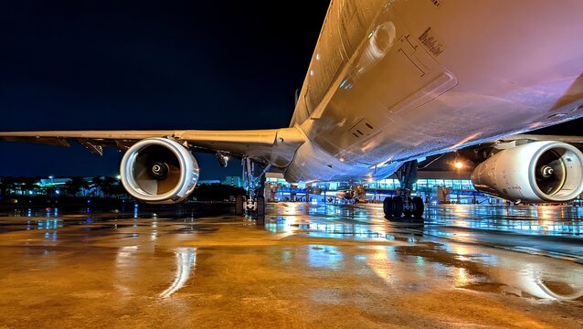 A large commercial aircraft parked on wet apron of Soekarno&ndash;Hatta International Airport at night, captured from a low angle showcasing its powerful engines, landing gear, and reflective ground lights