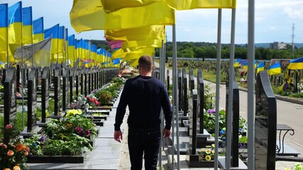 Mourning man go among countless graves of ukrainian soldiers with national flags at military cemetery in Kharkiv. Scene reflects personal sorrow and nation collective loss caused by russian aggression