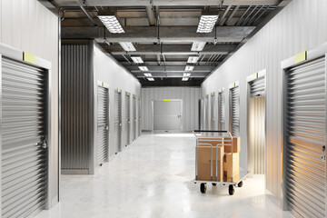 Clean, well-lit self-storage hallway with multiple metal roll-up doors, exposed ceiling pipes, and a cart loaded with cardboard boxes. Modern indoor storage facility. 3d image. © Grispb