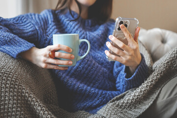 Female relaxing indoors in warm winter atmosphere, wearing knitted sweater and holding smartphone and mug. Soft cozy mood with seasonal comfort, warm textures and calm home during cold weather
