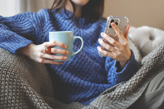 Female relaxing indoors in warm winter atmosphere, wearing knitted sweater and holding smartphone and mug. Soft cozy mood with seasonal comfort, warm textures and calm home during cold weather