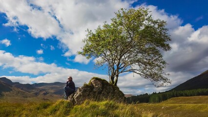 On a lovely day, a man sitting under a tree indulges in deep thought, after hiking through the highlands of Scotland in the UK.