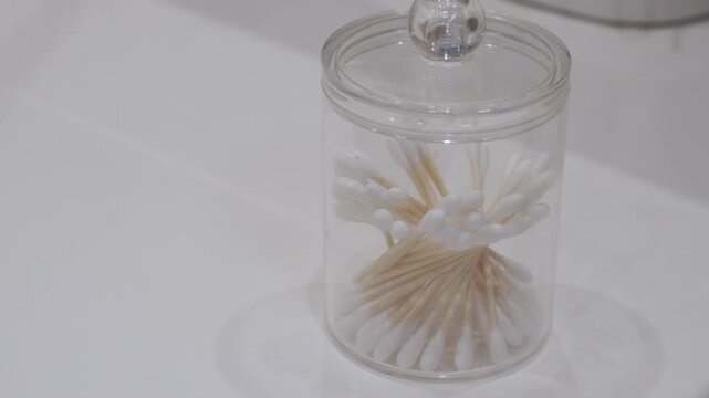 A clear plastic jar, filled with cotton swabs (Q-tips), is open as a hand finishes placing the lid. The action is completed with a subtle, dry plastic clicking sound.