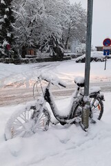 A snow-covered bike stands on the side of the road. In the background - a house, cars and trees covered with snow.