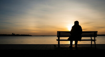 Obraz premium Contemplative silhouette watching the vibrant sunset over tranquil waters, embracing peace and solitude on a park bench at dusk