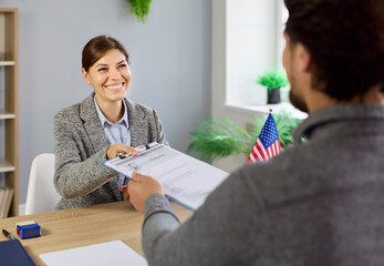 Consulate interview and approval of work visa documents in office with USA flag at desk. Happy female US embassy officer receiving documents to approve paperwork, resume verification from applicant
