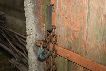 This close-up shot captures an old wooden door, featuring peeling orange paint, secured by a rusty chain and a chipped blue padlock. The door abuts a rough, aged concrete wall, creating an atmosphere 