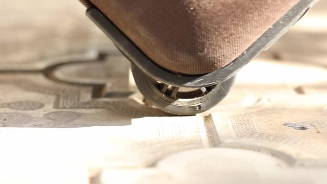 Extreme close-up of a brown luggage wheel rolling slowly across decorative, sunlit outdoor paving. This shot represents the themes of travel, journey, and movement.