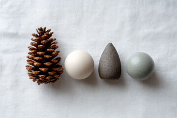 Natural pine cone and three decorative objects in neutral tones arranged in minimalist style on white fabric, evoking calm and simplicity