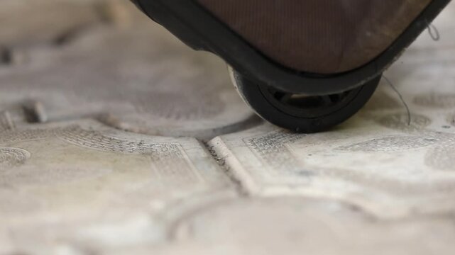Extreme close-up of a brown luggage wheel rolling slowly across decorative, sunlit outdoor paving. This shot represents the themes of travel, journey, and movement.
