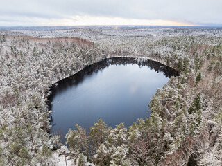 Wide aerial drone shot of Estonia's vast, snow-covered winter forest landscape with the dark, unfrozen Lake Vaike Kaksjarv.