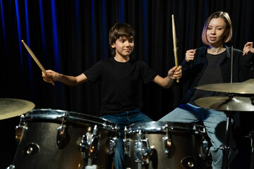 Fototapeta premium Young caucasian woman teaches a boy to play the drums in the studio on a black background. Music school student