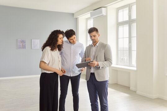 Young married couple man and woman standing with male professional realtor agent in empty new modern apartments discussing contract buying property. Real estate market and relocation concept.