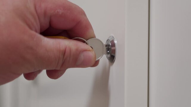 A person's hand is inserting a small silver metal key into a lock mechanism on a smooth white door or cabinet. The action is turning the key to lock or unlock the security device.