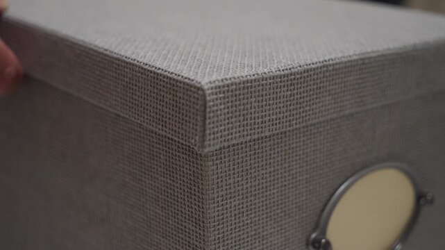 A close-up view of the corner of a woven-textured grey storage box with a metal-rimmed label holder is shown. The box is ready to be used for organizing.