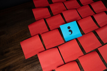 Single semi colon on blue sticky note in a grid pattern of red sticky notes on top of dark wood surface.  Your story isn't over.