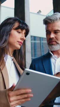 Close up of two business people, a man and a woman in formal attire, are standing outside a modern office building, discussing work-related topics using a digital tablet and a laptop