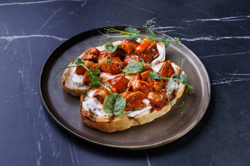Close-up of tomato bruschetta on artisan bread slices with fresh sliced tomatoes, parmesan cheese and herbs on blue plate, cooking moment. Food preparation process, bruschetta, sandwiches.