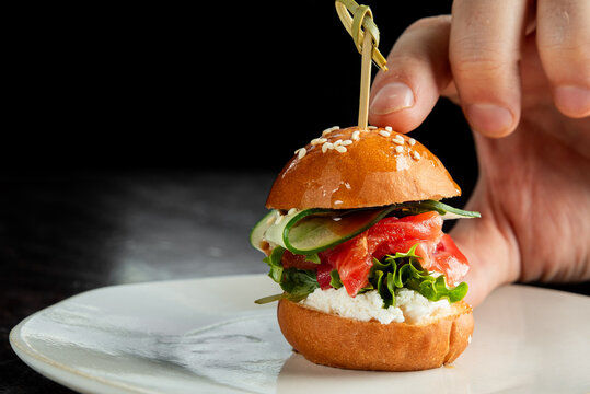 A hand holds a gourmet mini burger with salmon, cream cheese, tomato, and cucumber on a sesame seed bun, secured by a skewer on a white plate