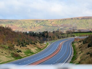 An elevated wide shot captures a modern, dual-carriageway highway curving through a rolling, semi-rural landscape under a pale, overcast sky. The road is flanked by grassy slopes and sparse trees