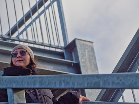 A low-angle, medium close-up of a woman standing on a bridge. She is leaning on the metal railing, wearing sunglasses, a beanie, and a scarf. She looks pensive or relaxed - Powered by Adobe