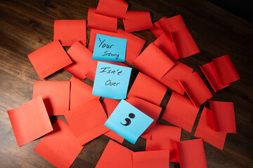 Text and single semi colon on blue sticky notes in a grid pattern of red sticky notes on top of dark wood surface.  Your story isn't over.