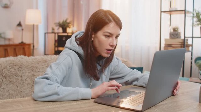 Happy young Caucasian woman browsing online store on laptop suddenly exclaims Wow after finding incredible discount celebrating online victory or good news. Excited girl sitting on sofa at table.