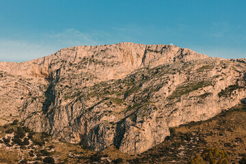 Wide view of a huge, striated rock mountain face with sparse vegetation, showing the small, visible moon near the upper ridge against a blue sky.