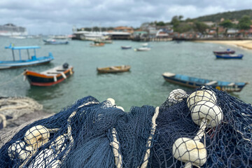 Close-up of a blue fishing net with white floats in the foreground, overlooking a small harbor on a cloudy day. 