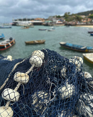 Close-up of a blue fishing net with white floats in the foreground, overlooking a small harbor on a cloudy day. 