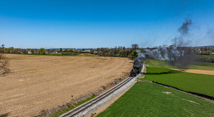 A vintage steam locomotive chugs along railway tracks surrounded by lush green fields and a clear blue sky. The train emits a plume of smoke as it journeys through the countryside.