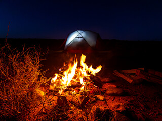 Campfire glowing at night with a lit tent in the Escalante desert
