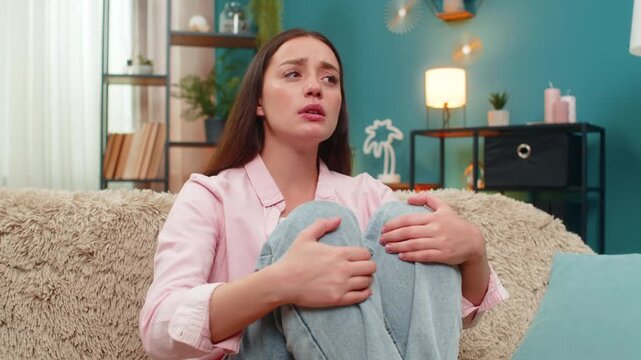 Young woman sits on home sofa hugging legs close feeling depression and stress after breakup job loss and bankruptcy. Caucasian girl stares downward apathetic on bad day seeking comfort in silence
