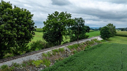 A railway track winds through lush green fields with scattered trees under a cloudy sky. The scene captures the serenity of rural life, showcasing nature and rail infrastructure.