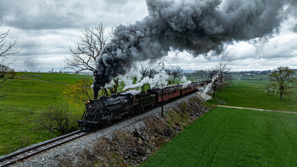 A vintage steam train chugs along railway tracks, billowing thick smoke against the backdrop of lush green fields and overcast skies. Trees line the path as it moves through the serene countryside.