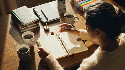 Woman sitting at a wooden desk with notebooks and coffee cups, writing in a planner with a pen.