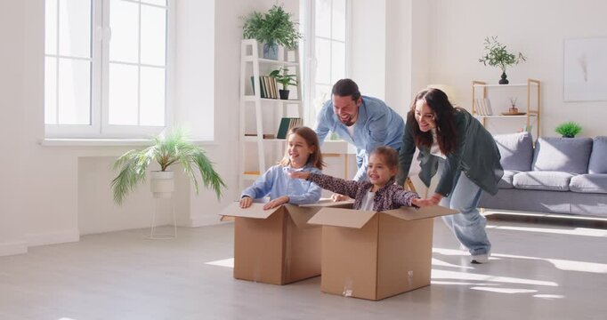 Parents engage children in playful activity by rolling them around living room in cardboard boxes. Brother and sister sit in boxes while mother and father playfully push them across living room floor.