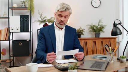 Middle-aged businessman at home office reviews budget today on laptop using calculator scanning receipts worried. Freelancer guy at table inputs figures adjusts expenses forecasting cash flow balance. - Powered by Adobe