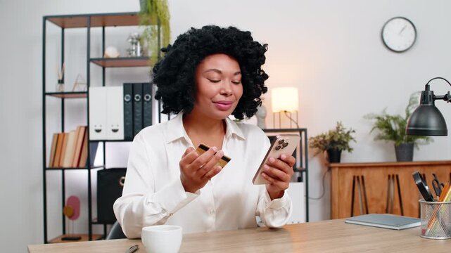 African American businesswoman at home office smiles using smartphone and credit card completing online payment. Freelancer girl at table celebrates purchase relaxing waiting delivery happy emotion