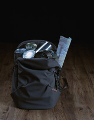 A backpack is full of cans and a map. The backpack is on a wooden floor. Ready for the budget trip