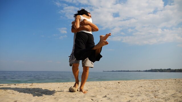 Happy couple having fun together on seashore at summer vacation. Man taking woman in air and turning around at coastline. Carefree lovers spending time at sandy beach. Concept of love. Slow motion