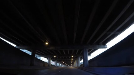 POV drive under a huge, dimly lit concrete overpass at high speed in the city. The darkness emphasizes the repetitive structure, hinting at the urgency associated with a police siren.