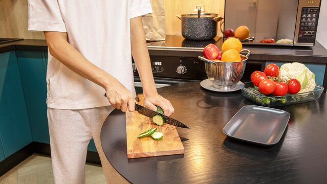 Person cutting fresh cucumbers on a wooden board surrounded by vegetables and fruits on the kitchen table. Healthy salad preparation and clean eating lifestyle at home.