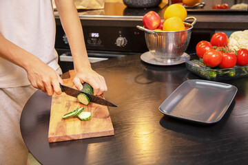 Person cutting fresh cucumbers on a wooden board surrounded by vegetables and fruits on the kitchen table. Healthy salad preparation and clean eating lifestyle at home.
