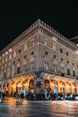 Naklejka premium Exterior corner view of a large, well-lit historic building on a cobblestone street at night with people gathered near the ground floor.