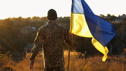 Soldier of ukrainian army going with lifting blue-yellow banner at sunset. Young male military in uniform walks with waving flag of Ukraine at countryside. Victory against russian aggression concept