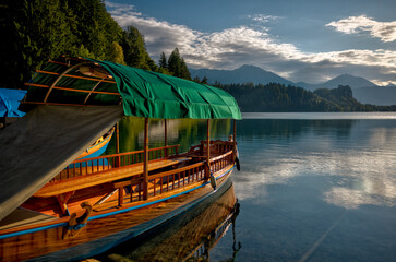 Traditional wooden pletna boats docked along the calm waters of Lake Bled, Slovenia, surrounded by lush greenery and the Julian Alps in the distance on a serene summer morning.