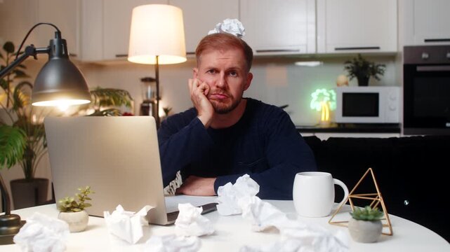 Young man at night home writes ideas for project faces creative block feels stress mood. Caucasian guy on sofa at table looks at laptop sees crumpled paper notes does critique tries to plan work hard.