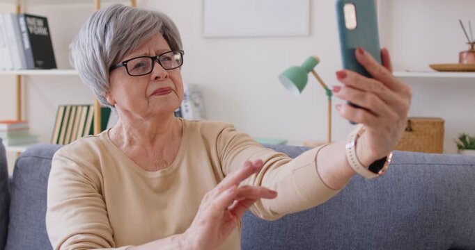 Senior woman holding smartphone, trying to make selfie portrait, adjusting camera, wearing eyeglasses, posing on sofa with modern gadget, preparing for video conference. Technology concept