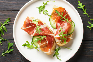 Open sandwiches with cream cheese, prosciutto, cucumber and arugula on white plate.